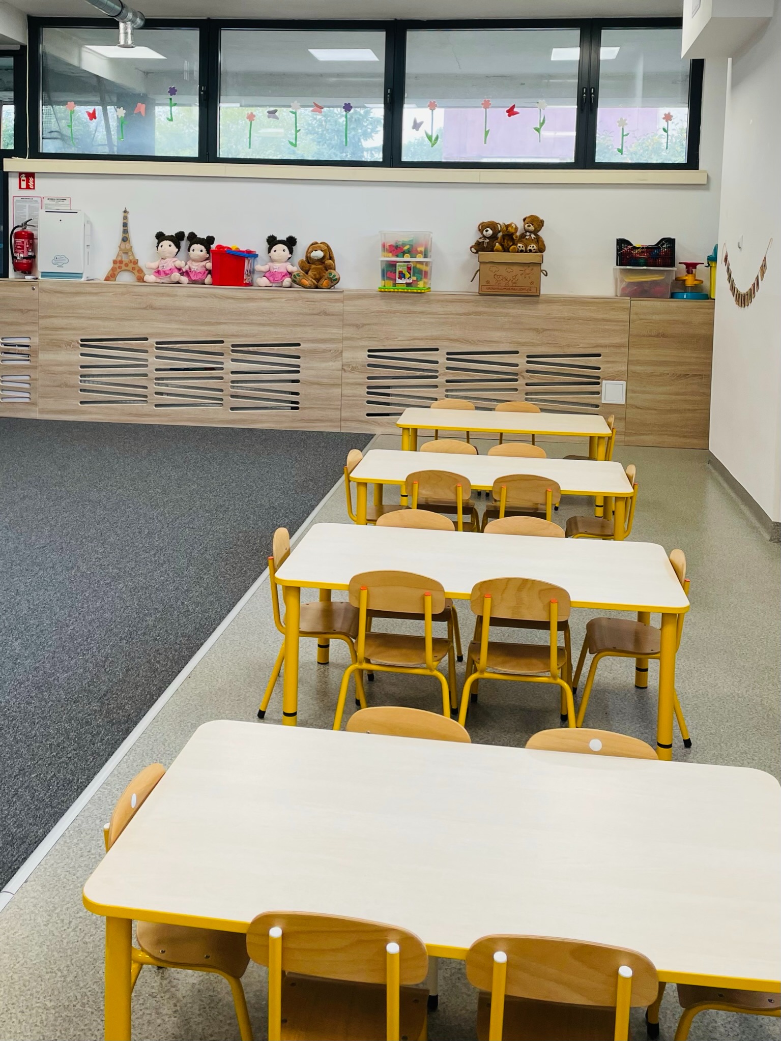 Sala zabaw z rzędami żółtych stolików i krzeseł, na szafce zabawki, dekoracje okienne z motywem kwiatów i motyli.