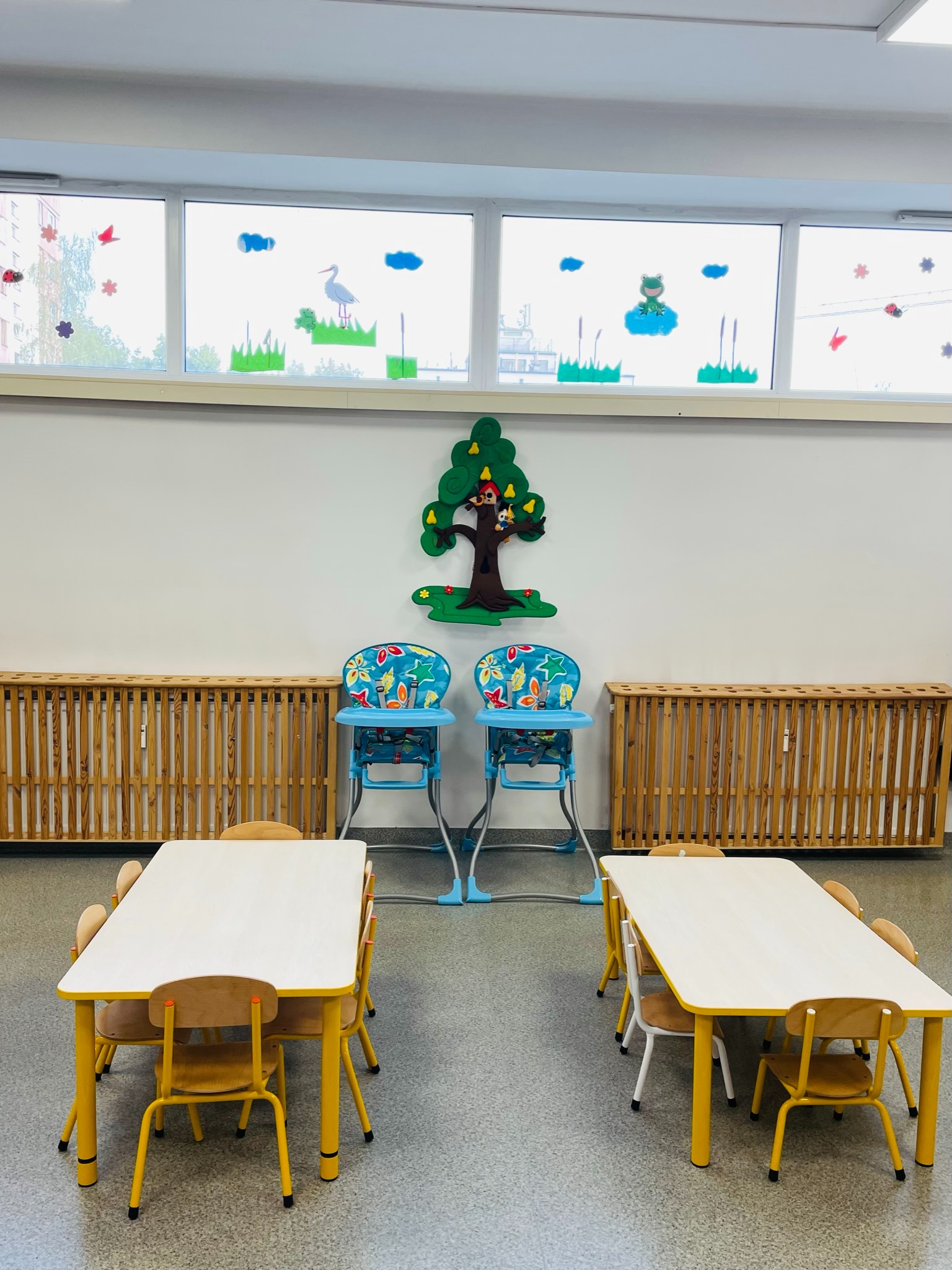 Jasne wnętrze sali zabaw z dwoma stolikami z żółtymi nogami i krzesełkami, dwoma krzesełkami do karmienia, dekoracjami okiennymi z motywami zwierząt i roślin oraz drewnianym grzejnikiem.