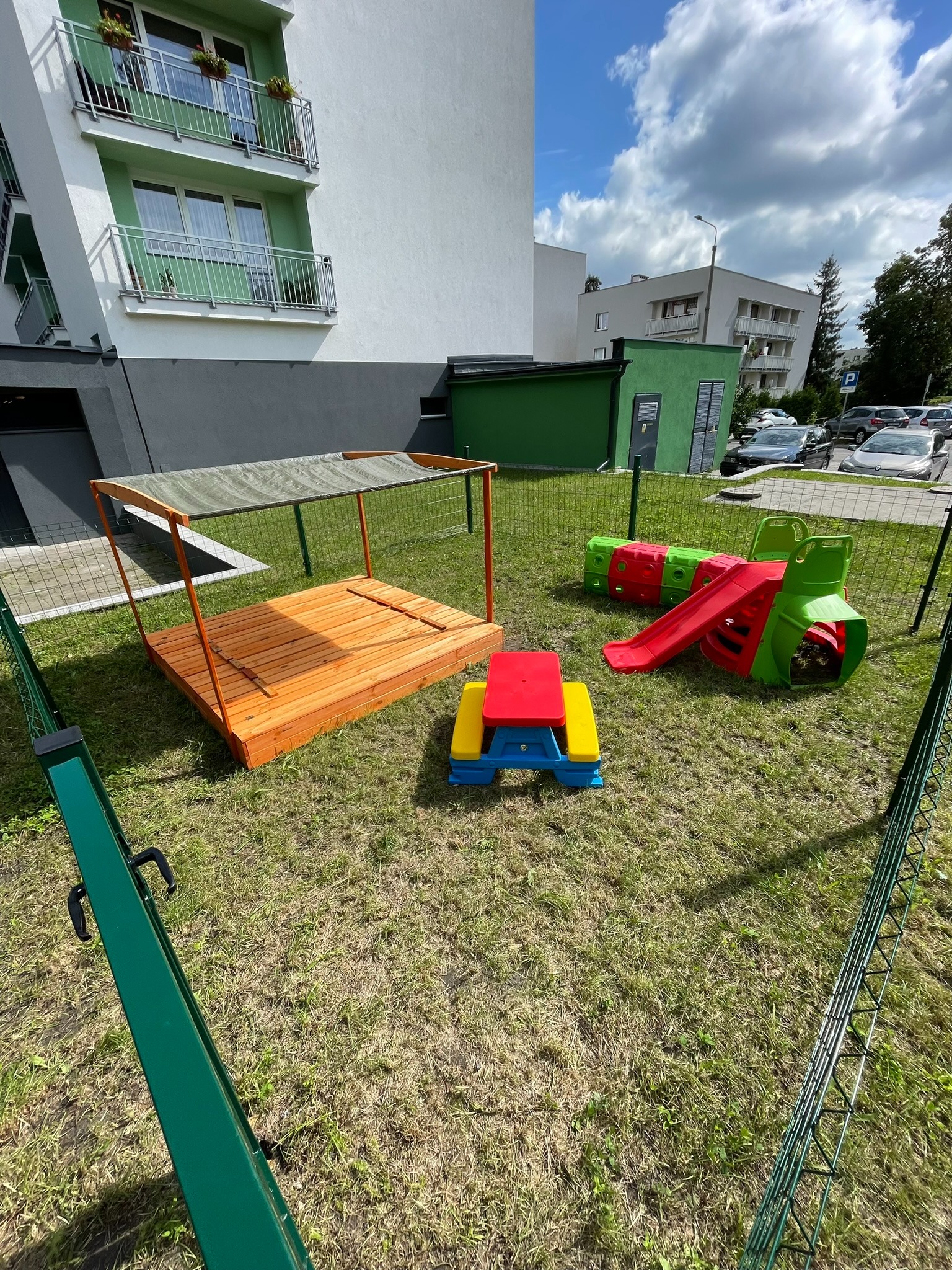 Plac zabaw na trawniku przed budynkiem mieszkalnym: piaskownica z zadaszeniem, kolorowa zjeżdżalnia-tunel i stolik piknikowy dla dzieci.