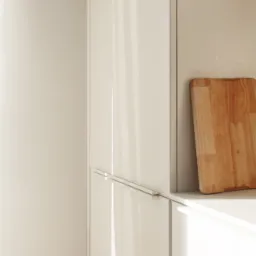 Minimalistyczna kuchnia z białymi, lakierowanymi frontami szafek i drewnianą deską do krojenia opartą o ścianę, oświetlona naturalnym światłem.