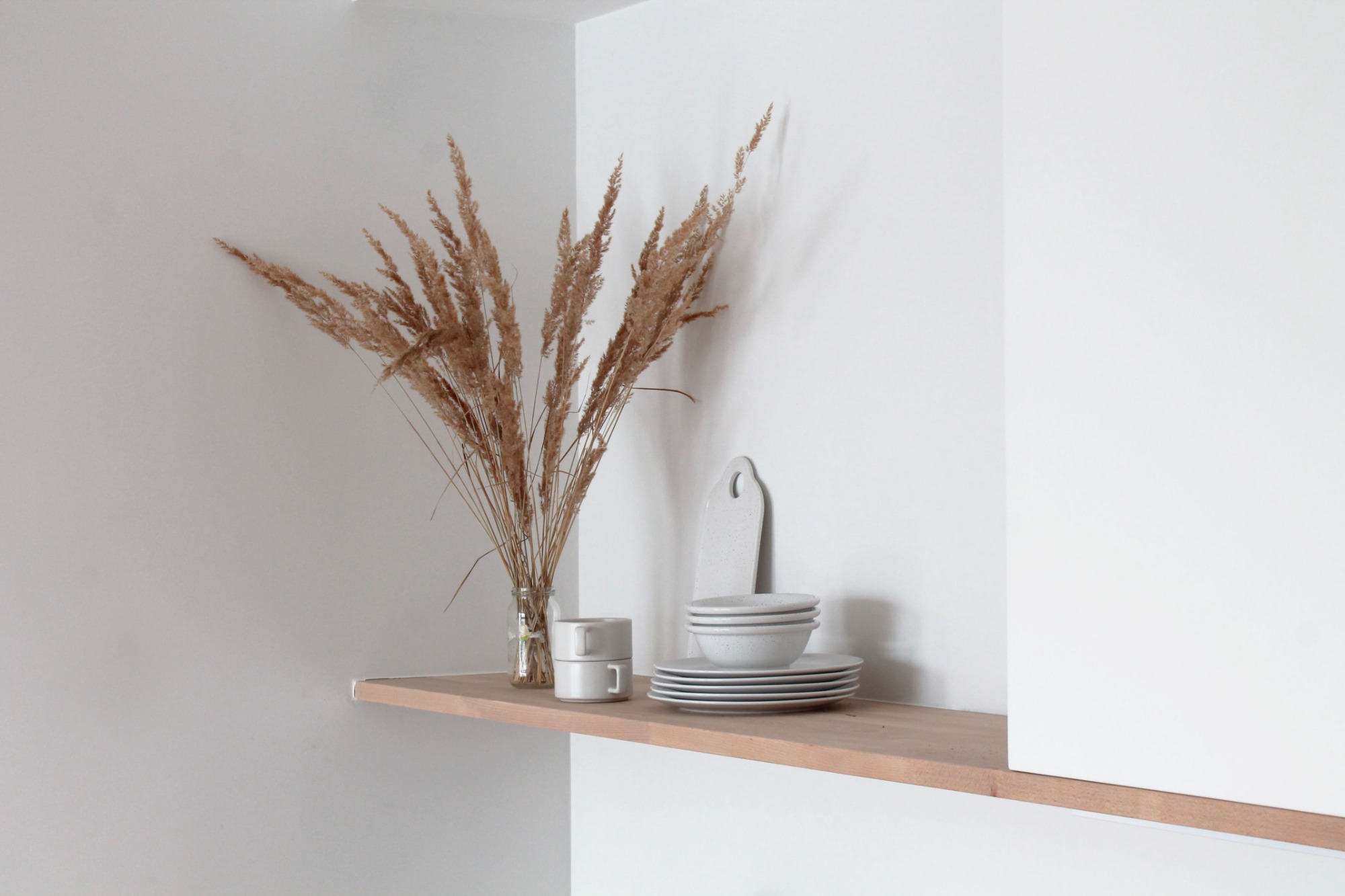 Minimalistyczna aranżacja półki z drewnianym blatem: szklany wazon z suszonymi trawami, dwie ceramiczne filiżanki w stosie, zestaw naczyń i deska do krojenia w odcieniach bieli