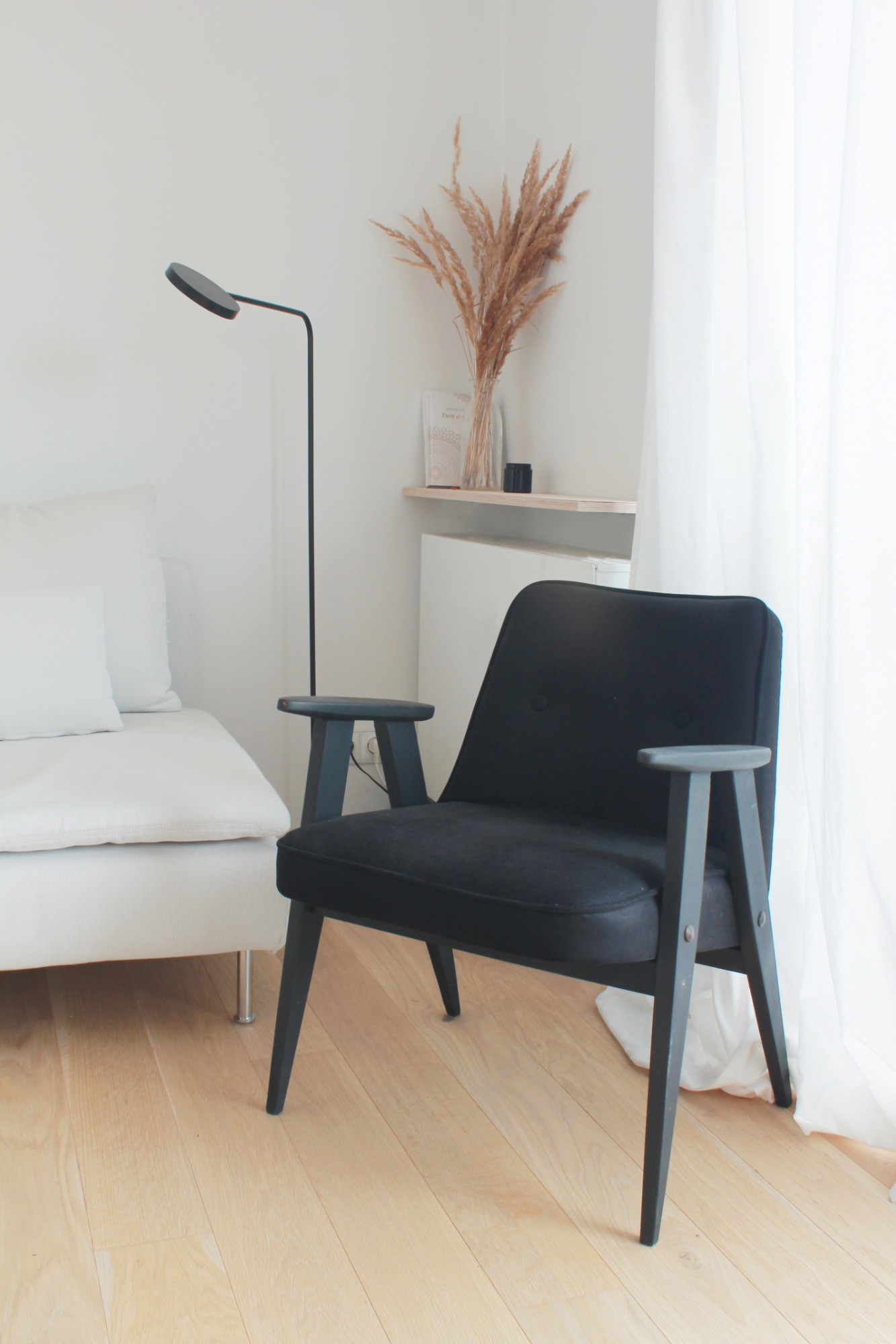 Minimalistyczne wnętrze: czarny fotel na drewnianej podłodze, obok biała sofa, lampa podłogowa i dekoracja z suszonych traw w szklanym wazonie na półce.