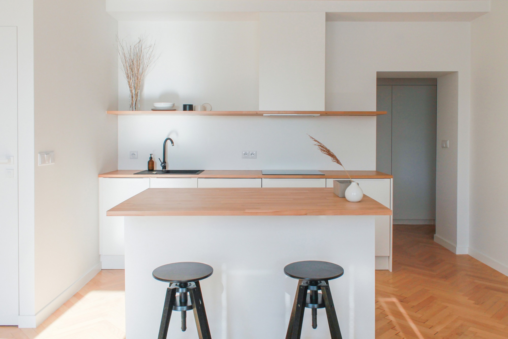 Minimalistyczna kuchnia z białymi szafkami, drewnianym blatem, czarną armaturą, drewnianą podłogą w jodełkę i dwoma czarnymi stołkami barowymi