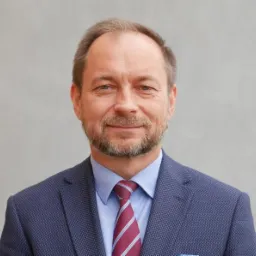 Portret mężczyzny w średnim wieku, w granatowej marynarce w drobną kratę i bordowym krawacie w białe paski, uśmiechającego się delikatnie na tle szarej ściany.