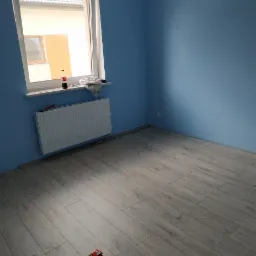 Pomalowane na niebiesko ściany pokoju z ułożonymi panelami podłogowymi, widoczny grzejnik pod oknem, butelka coli i miarka na podłodze.