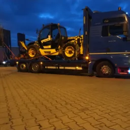 Niebieska ciężarówka MAN z żółtą ładowarką teleskopową New Holland na niskopodwoziowej platformie, sfotografowana na parkingu z kostki brukowej o zmierzchu.