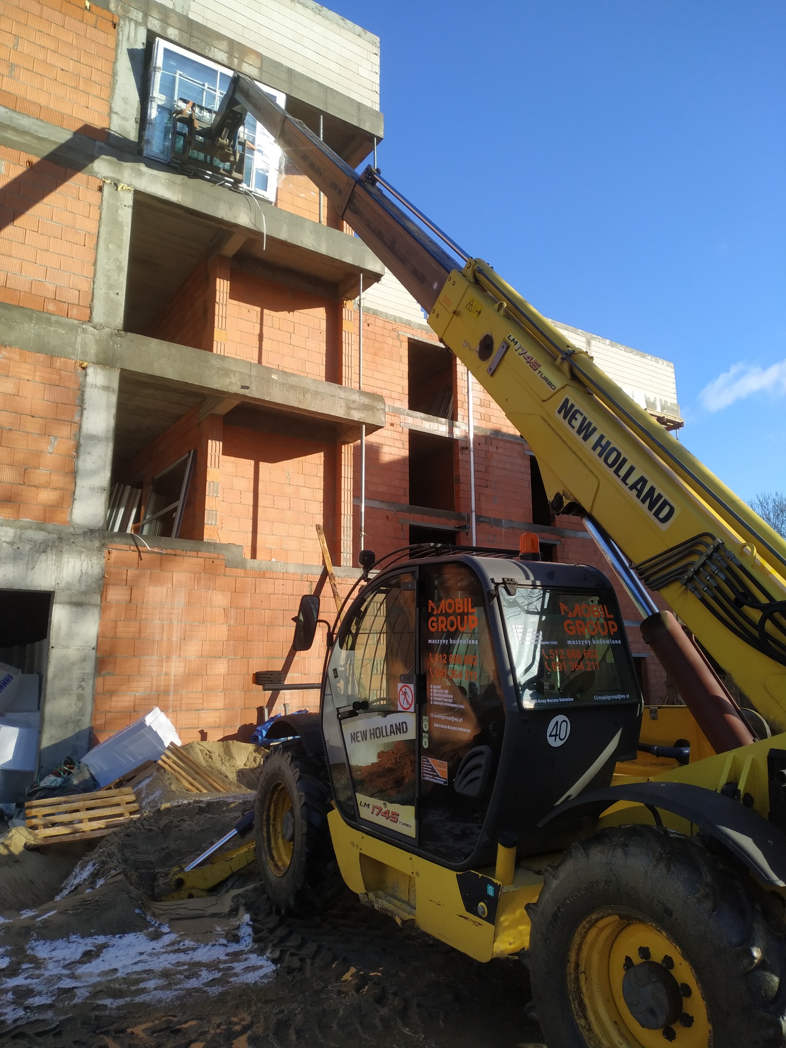 Żółta ładowarka teleskopowa New Holland podnosi okno na placu budowy, widoczne ceglane ściany w stanie surowym i materiały budowlane obok, w tle błękitne niebo.