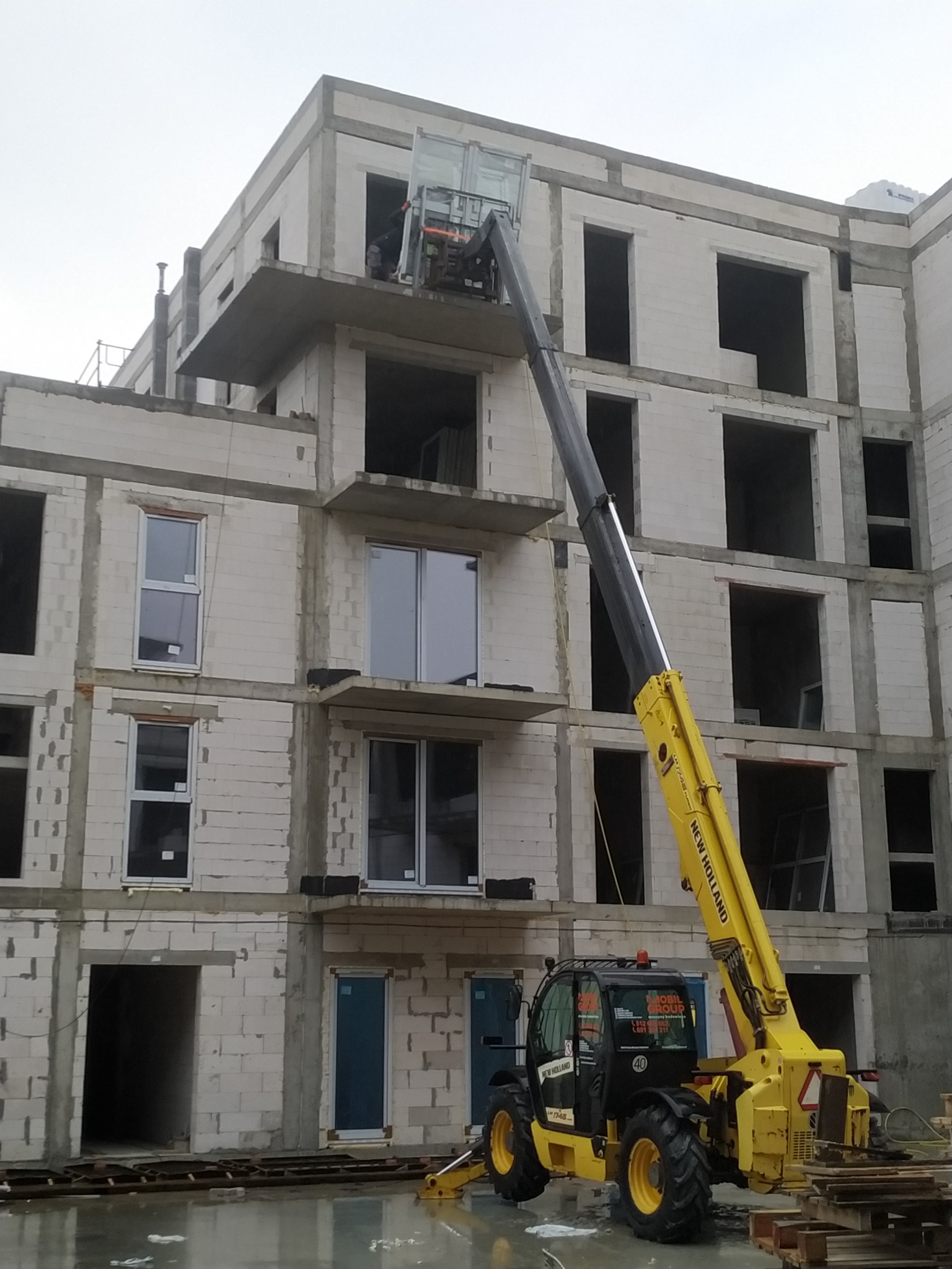 Żółty dźwig teleskopowy marki New Holland podnosi okno na wyższe piętro budynku w stanie surowym w Wyszkowie. Na ziemi widoczne kałuże po deszczu i palety.