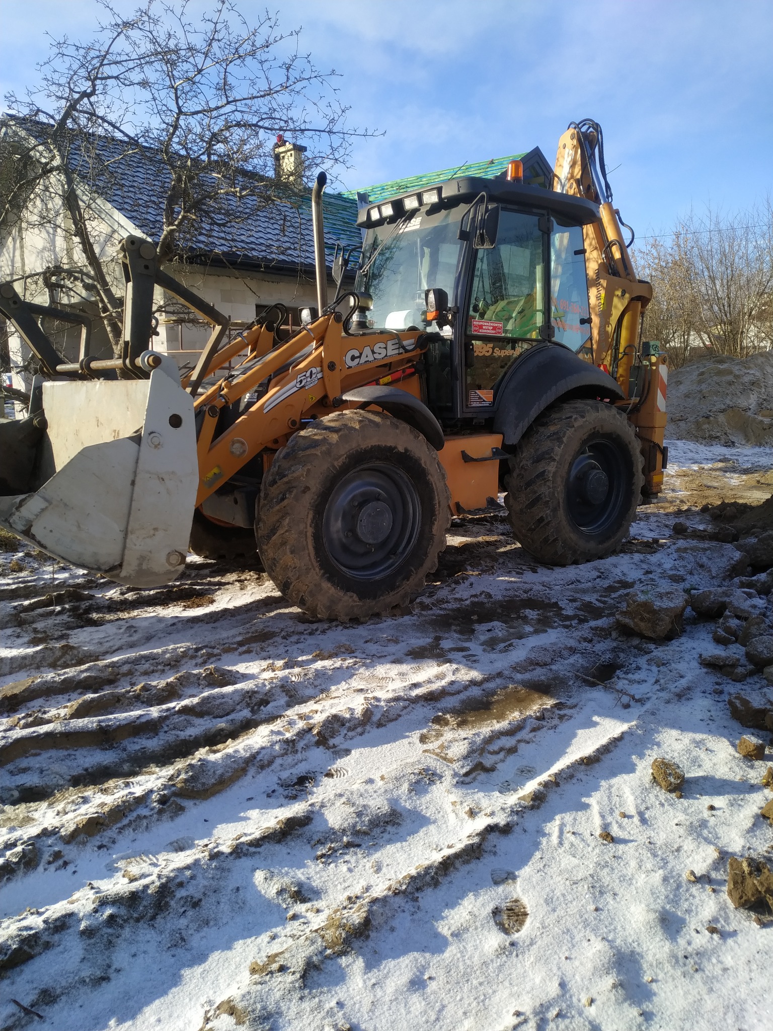 Pomarańczowa koparko-ładowarka CASE 580 Super N na placu budowy w zimowy dzień, widoczne ślady opon w śniegu i częściowo odśnieżony teren wokół.