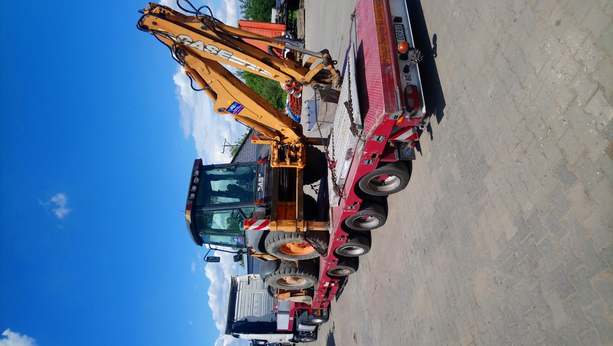 Żółta koparko-ładowarka Case na czerwonej niskopodwoziowej przyczepie transportowa, widok z boku na tle błękitnego nieba z chmurami.