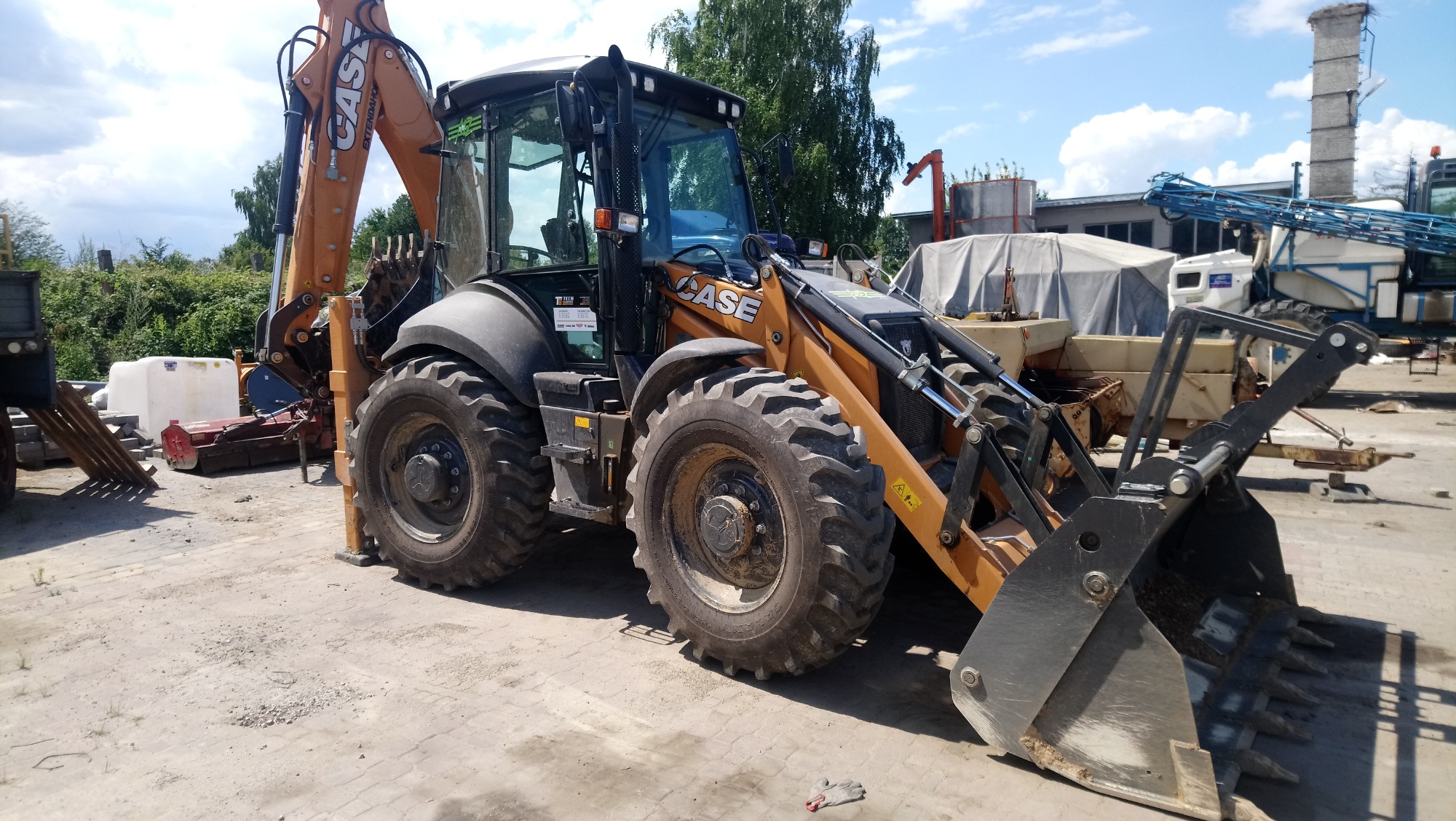 Pomarańczowo-czarna koparko-ładowarka marki Case stoi na placu budowy, z widocznymi śladami użytkowania na oponach i łyżce. W tle inne maszyny budowlane i zielona roślinność.