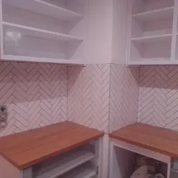 Ściana wyłożona jasnymi płytkami ułożonymi w jodełkę, nad drewnianym blatem i białymi szafkami wiszącymi, widoczny fragment instalacji elektrycznej.