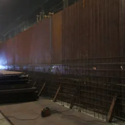 Spawanie konstrukcji stalowej zbrojonej siatką na tle dużej hali produkcyjnej, widoczny spawacz i sprzęt spawalniczy.