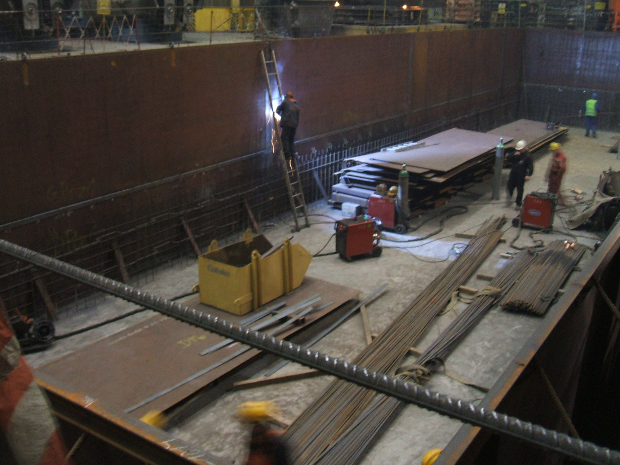 Spawanie metalowych płyt w dużym, przemysłowym zbiorniku; spawacz na drabinie łączy elementy, widoczne stosy blach, butle z gazem i sprzęt spawalniczy.