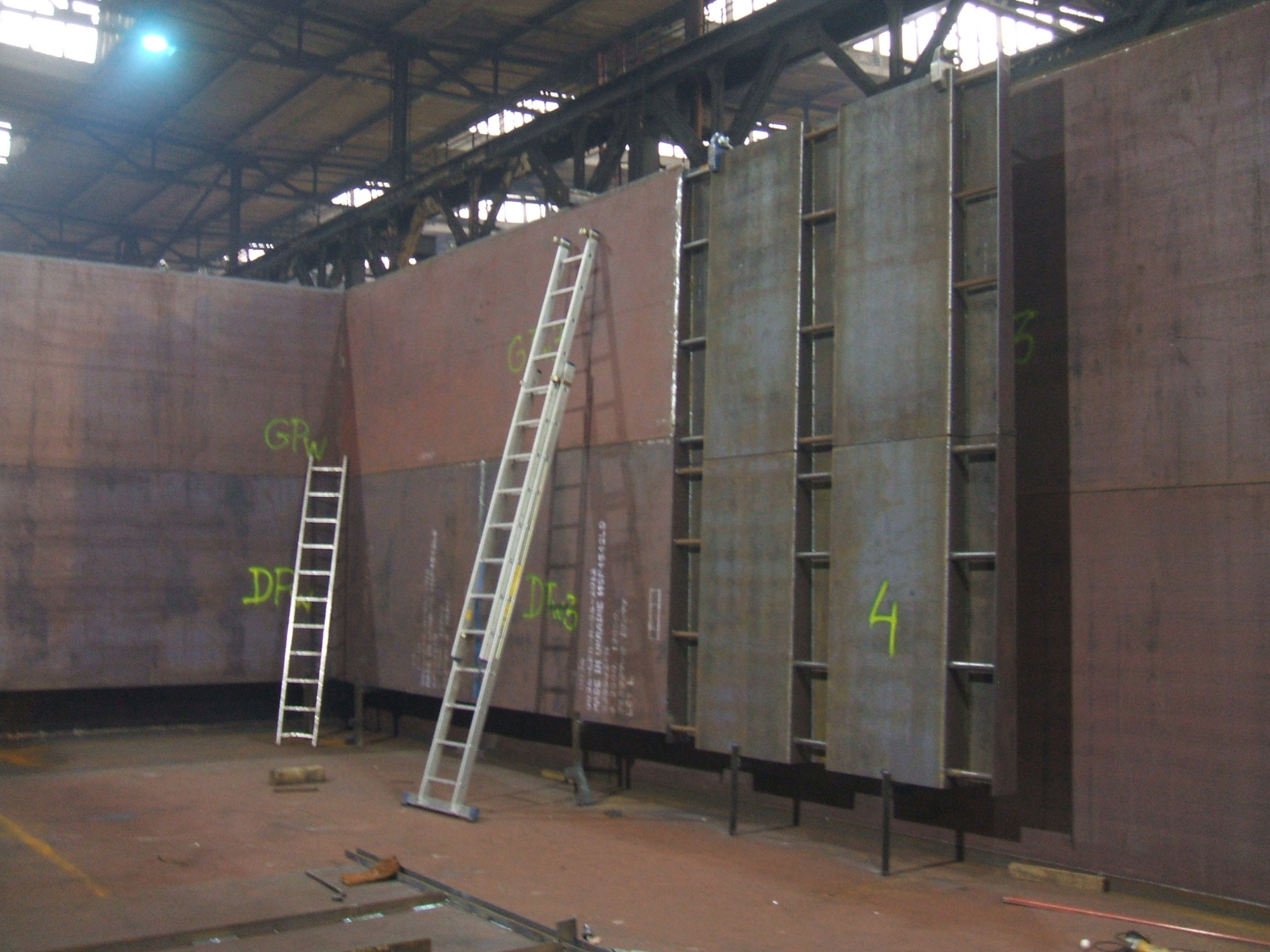 Wnętrze hali produkcyjnej z dwoma drabinami opartymi o pionową konstrukcję z płyt stalowych, niektóre z numerami, widoczne elementy spawane i ślady po obróbce.