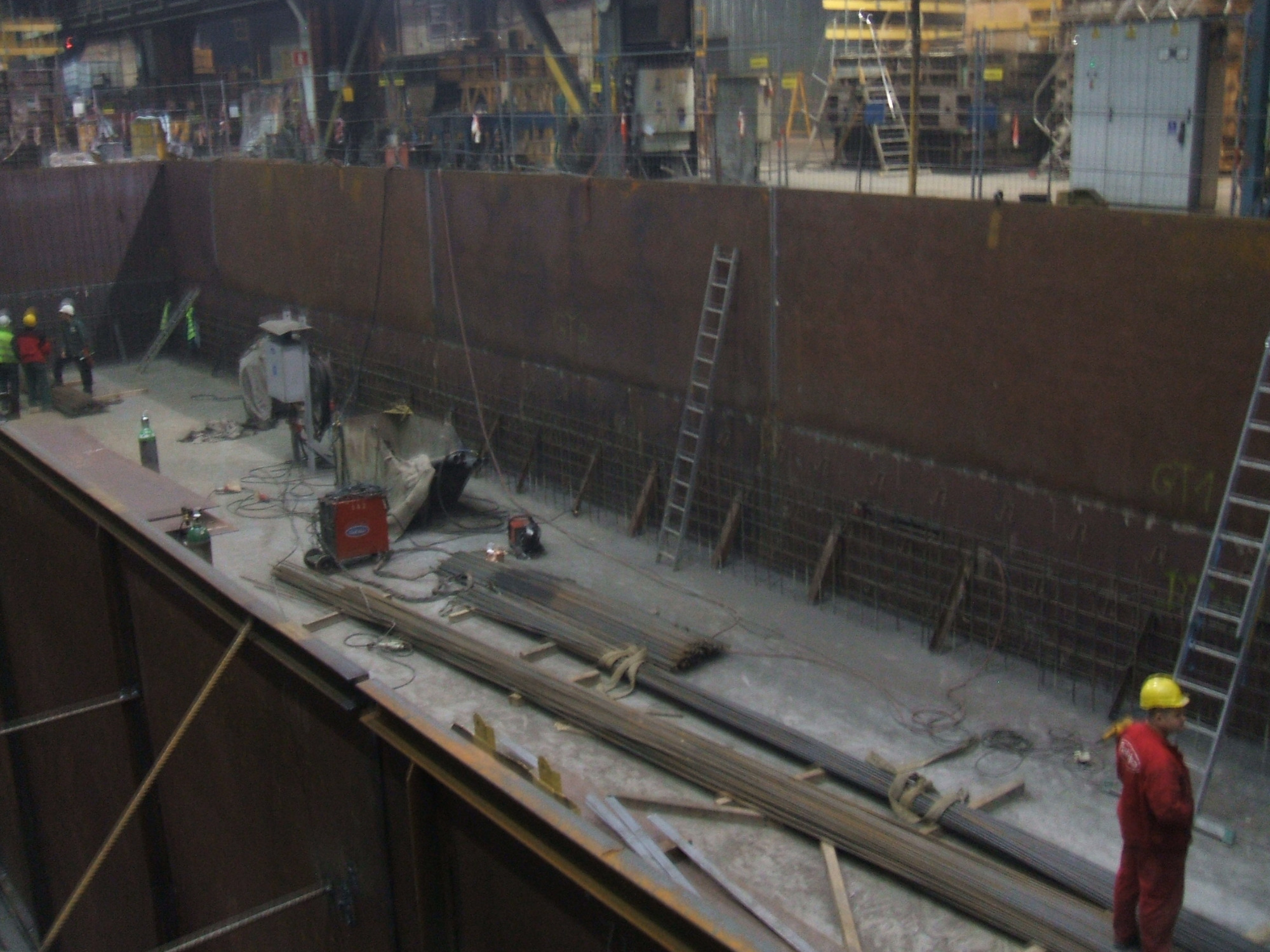 Wnętrze hali produkcyjnej z konstrukcją stalową, widoczne elementy balustrady nierdzewnej w trakcie montażu, spawarka, pracownicy w kaskach i kombinezonach, drabiny