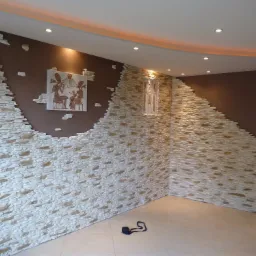 Fragment ściany w pomieszczeniu z dekoracyjnym wykończeniem z kamienia imitującego piaskowiec, przerywanym na brązowej ścianie z obrazkiem w stylu egipskim i małą figurką, beżowa podłoga...