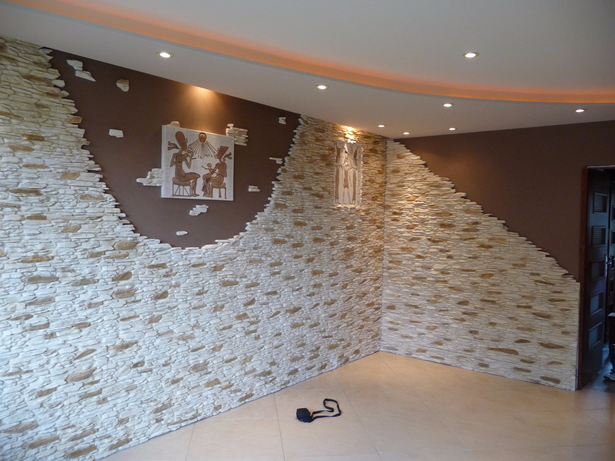 Fragment ściany w pomieszczeniu z dekoracyjnym wykończeniem z kamienia imitującego piaskowiec, przerywanym na brązowej ścianie z obrazkiem w stylu egipskim i małą figurką, beżowa podłoga...