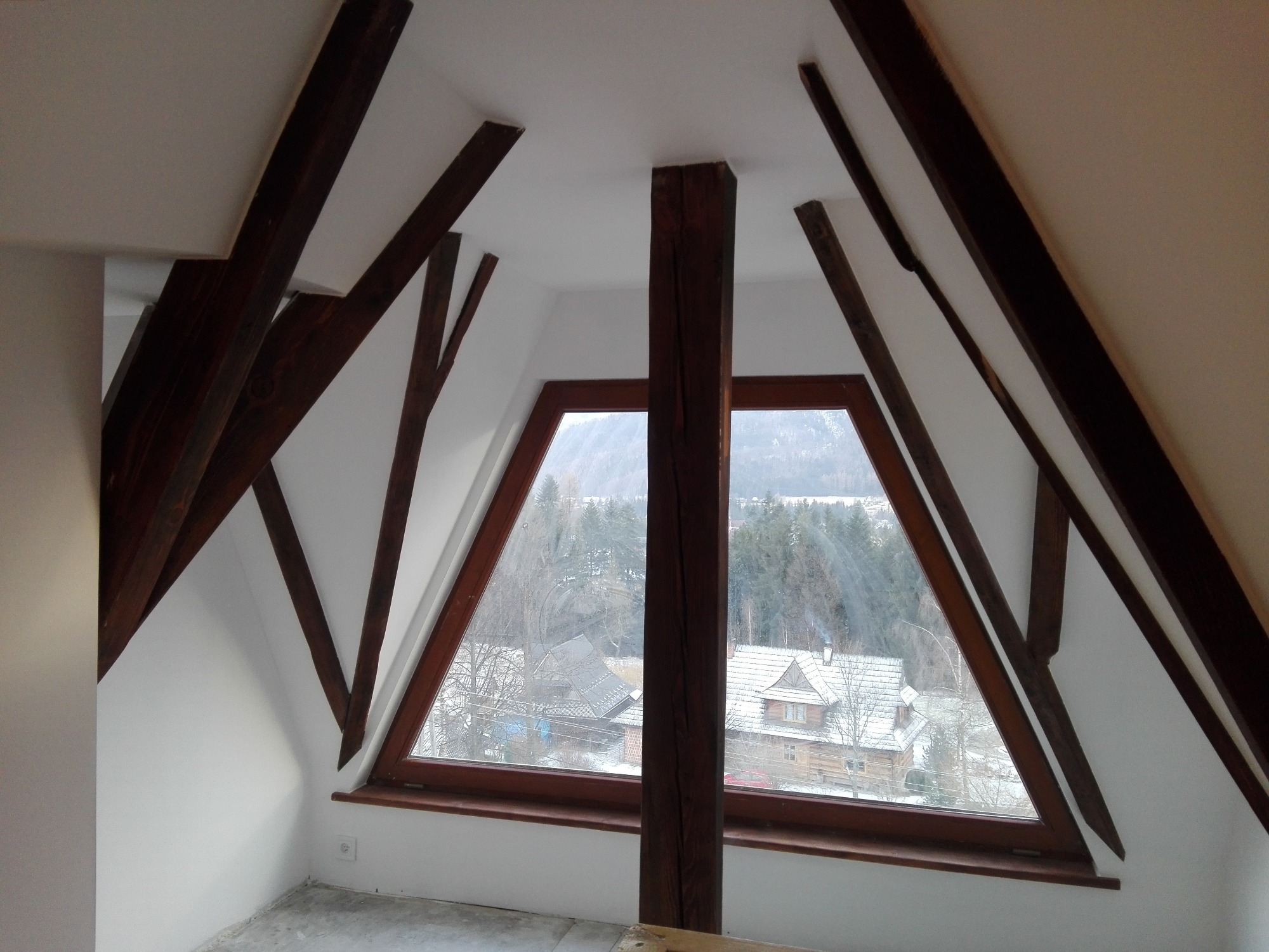Poddasze z drewnianą konstrukcją dachu i trójkątnym oknem z widokiem na zimowy krajobraz z domami.