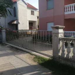 Nowoczesna, metalowa brama wjazdowa z pionowymi prętami, osadzona w betonowych słupkach z dekoracyjną balustradą, na tle dwóch budynków mieszkalnych w ciepłym świetle popołudniowym.