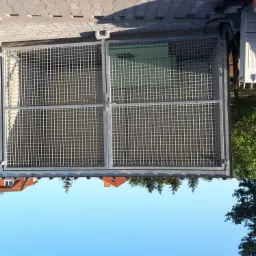 Metalowa krata zamykająca wnękę, widoczna z zewnątrz, z fragmentami budynku, drzew i nieba w tle.