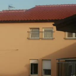 Fragment elewacji budynku z czerwonym dachem i balkonem z metalową konstrukcją, widoczne okna z roletami i bez, ujęcie z dołu w słoneczny dzień.