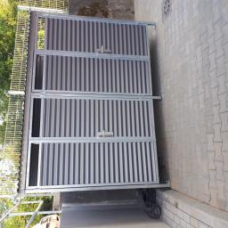 WELDER-SERWIS OLGA SIWIK - Metalowa konstrukcja z falistej blachy, prawdopodobnie boks na śmietniki, z widocznym zamkiem i fragmentem rynny, na tle kostki brukowej i zieleni.