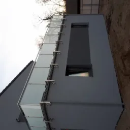 Nowoczesny budynek z szarą elewacją i balkonem z szkła i metalu, widok z dołu, surowy teren wokół budynku.