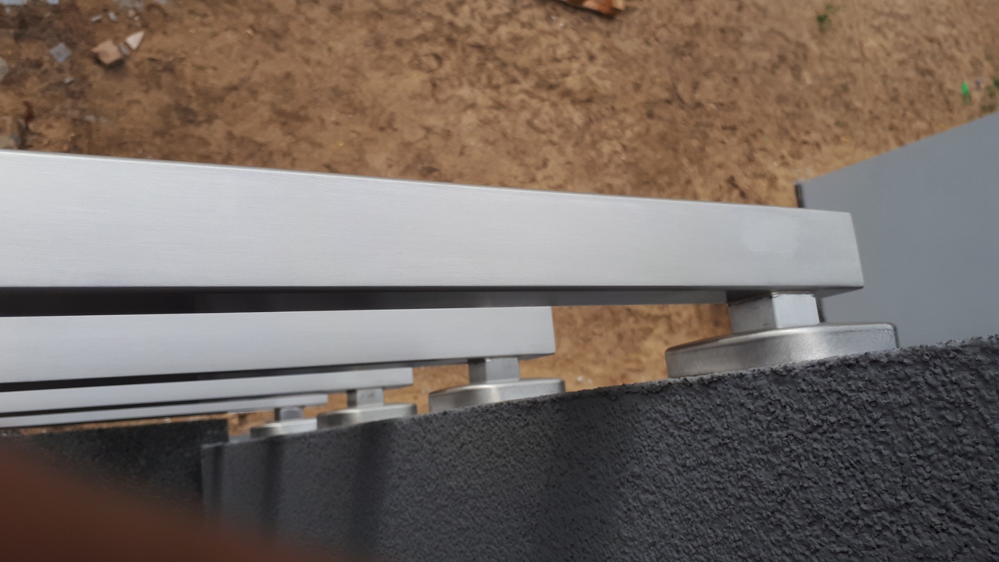 Szczegółowe ujęcie mocowania metalowej konstrukcji, prawdopodobnie bariery lub balustrady, do szarego, chropowatego muru w Zgorzelcu. Widoczne elementy montażowe i fragmenty poziomych profili.