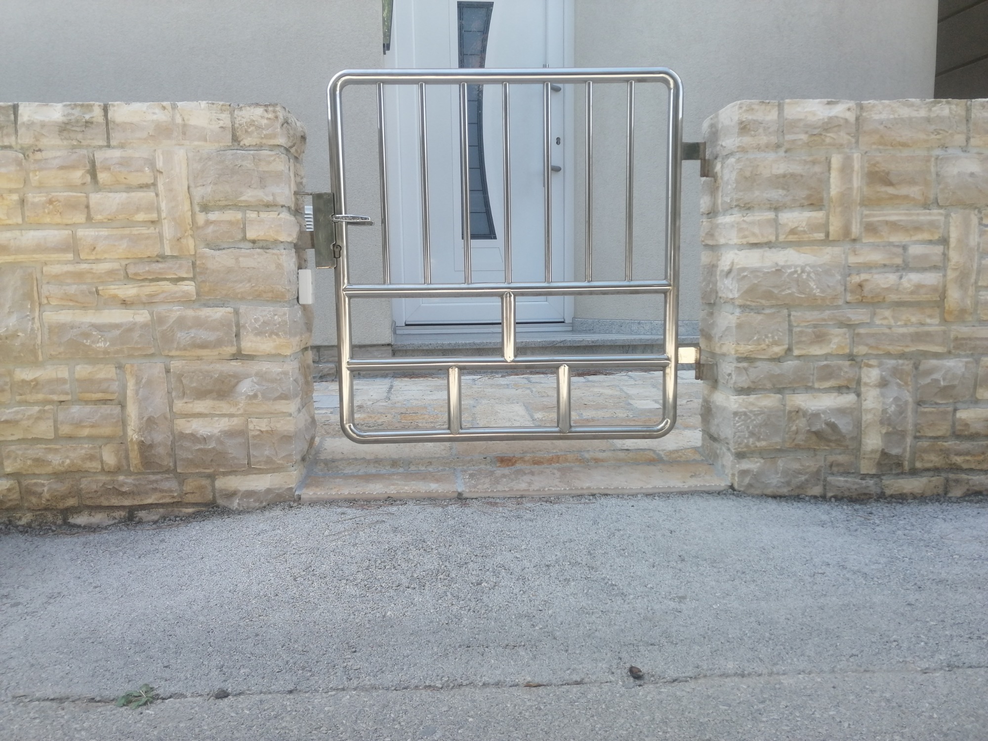 Brama ze stali nierdzewnej o minimalistycznym designie, zamontowana pomiędzy kamiennymi słupkami, prowadząca do drzwi wejściowych.