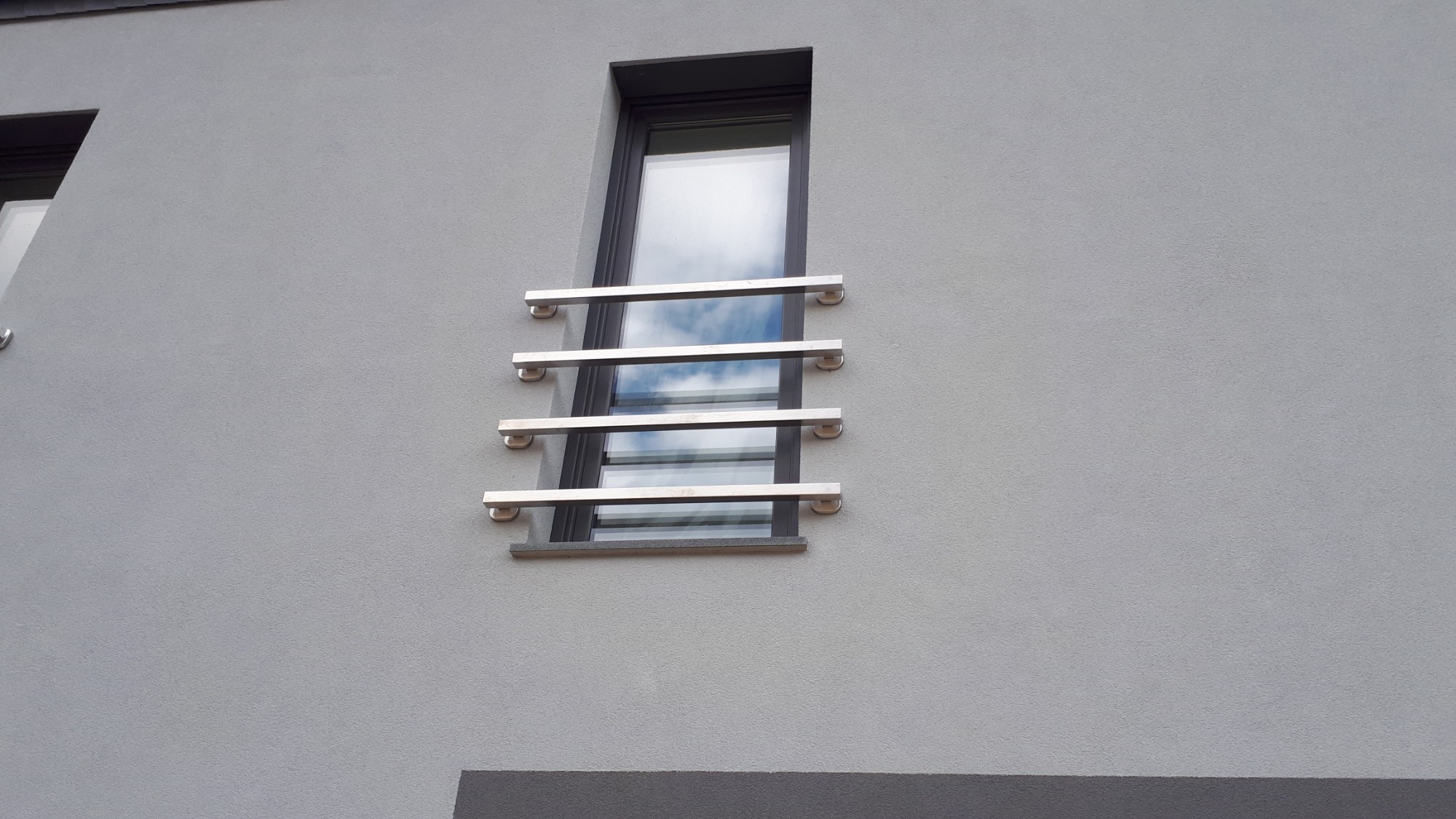 Nierdzewna balustrada okienna z poziomych rurek na szarej elewacji budynku, odbicie chmur w szybie.