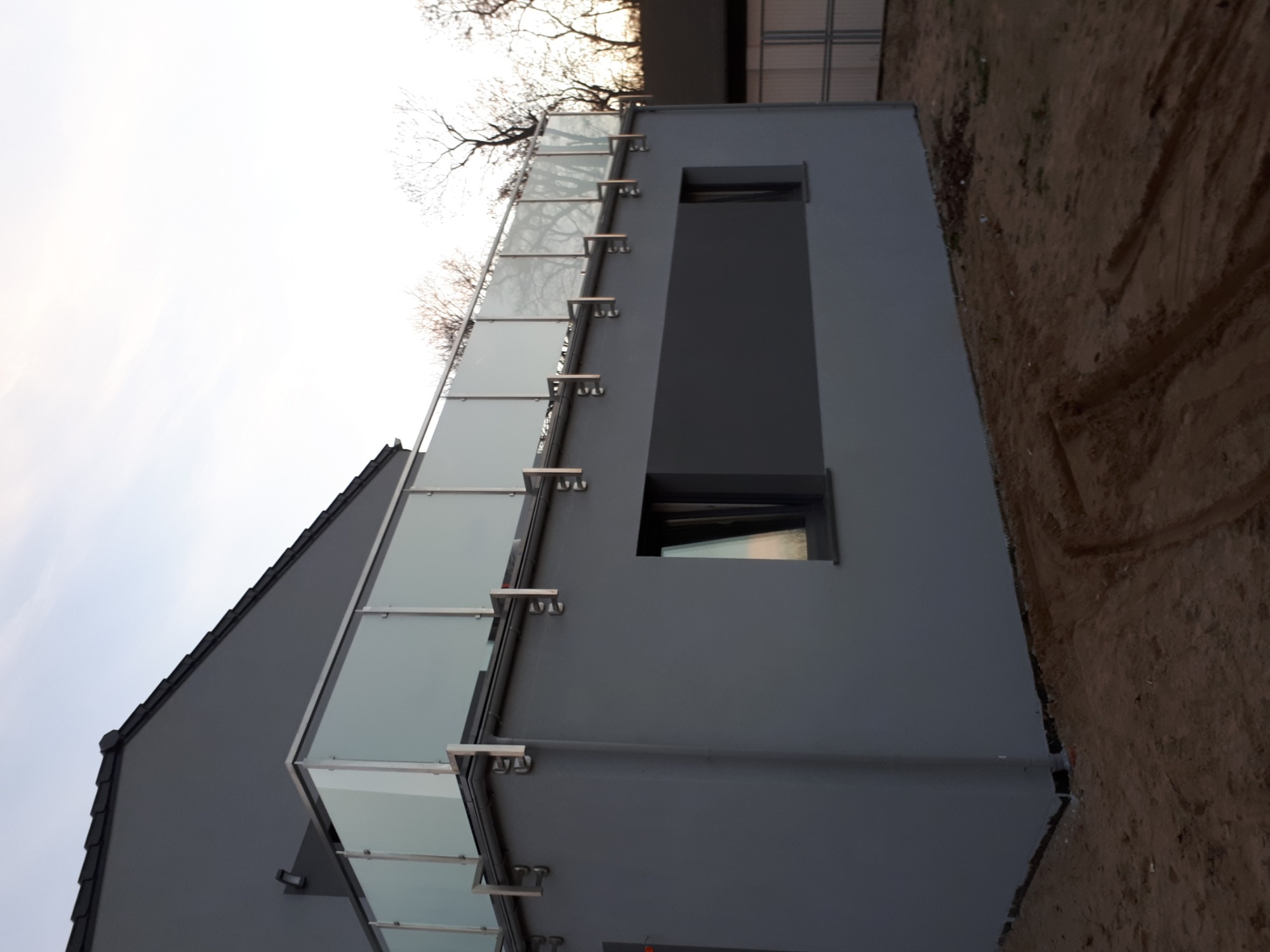 Nowoczesny budynek z szarą elewacją i balkonem z szkła i metalu, widok z dołu, surowy teren wokół budynku.