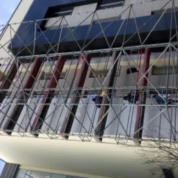 Budynek w trakcie ocieplania, widoczne rusztowania i pracownicy na wysokościach, fragment elewacji pokryty warstwą izolacyjną.