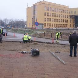 JM BUDINVEST - Układanie kostki brukowej na zewnątrz budynku, widoczni robotnicy w kamizelkach odblaskowych, sprzęt budowlany, generator prądu, oraz osoba nadzorująca prace. W tle budynek biurowy.