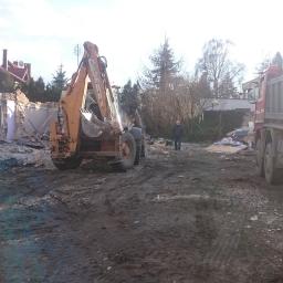 JM BUDINVEST - Pomarańczowa koparko-ładowarka Case na placu budowy podczas wyburzania ściany budynku, z gruzem, stojącym mężczyzną i ciężarówką w tle.