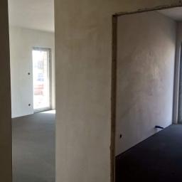 JM BUDINVEST - Wnętrze w stanie deweloperskim: surowe ściany z widocznymi śladami tynkowania, betonowa wylewka podłogowa, okno z widokiem na plac budowy, przygotowane otwory pod instalacje.