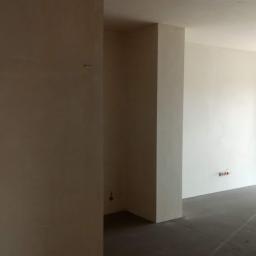 JM BUDINVEST - Wnętrze w trakcie prac wykończeniowych: ściany po tynkowaniu maszynowym, widoczne wystające kable elektryczne, surowa podłoga, widok na jasne drzwi balkonowe.