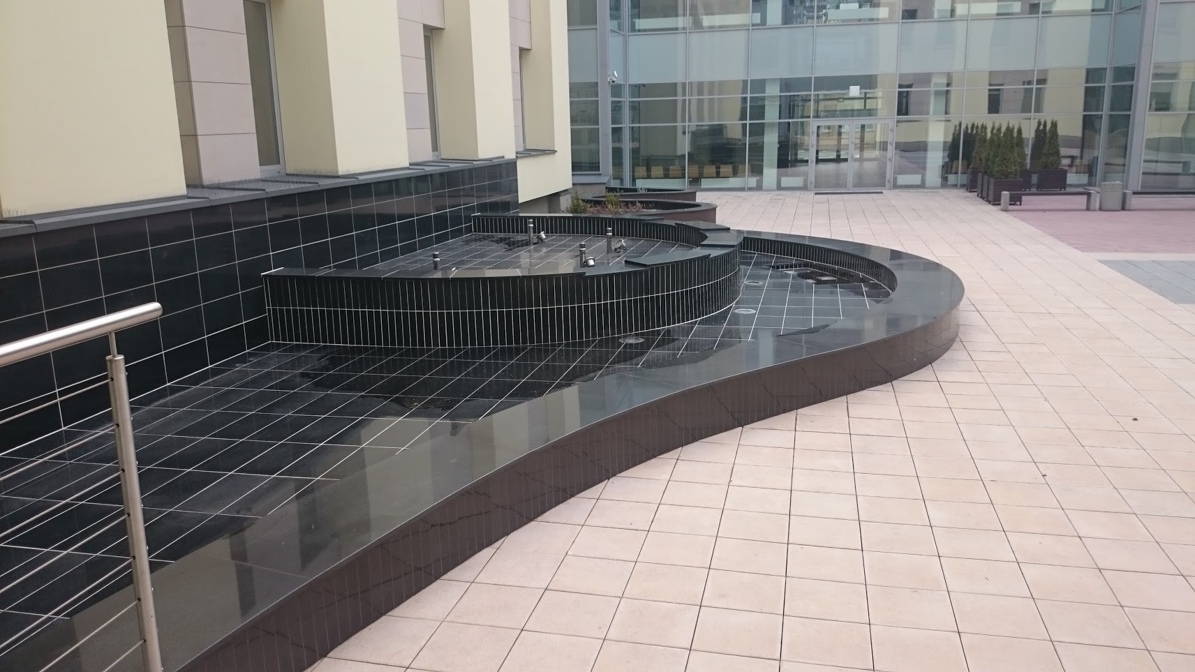 Czarna, kaskadowa fontanna z metalowymi kratkami w nowoczesnym otoczeniu biurowca, z widocznymi dyszami wodnymi i minimalistyczną zielenią.