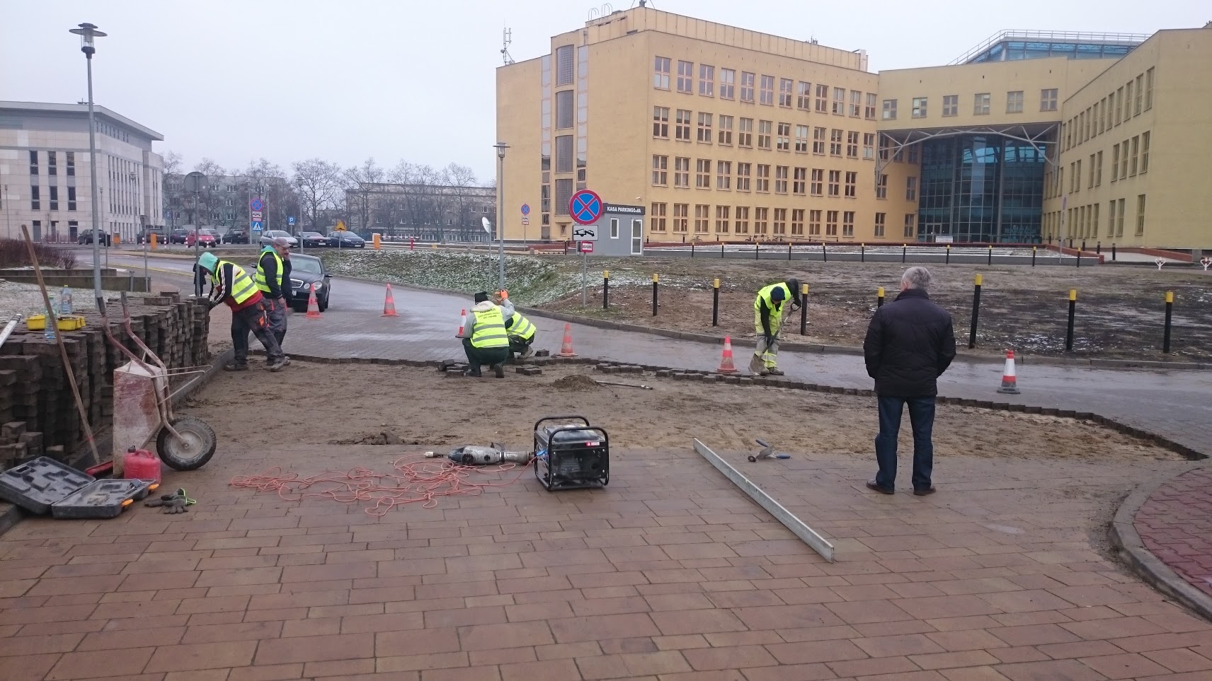 Układanie kostki brukowej na zewnątrz budynku, widoczni robotnicy w kamizelkach odblaskowych, sprzęt budowlany, generator prądu, oraz osoba nadzorująca prace. W tle budynek biurowy.