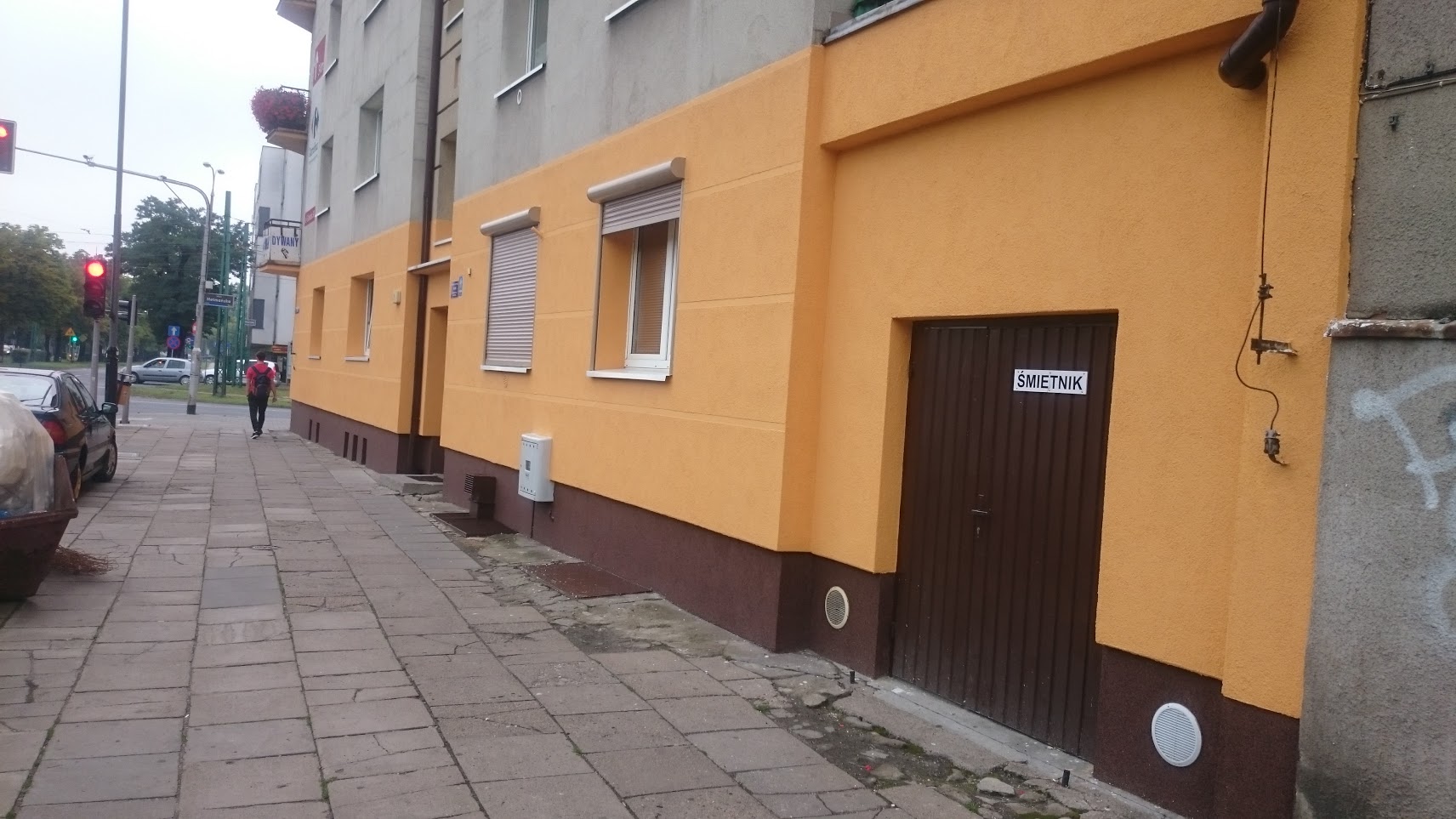 Budynek z ocieploną i pomalowaną na żółto elewacją, widoczne okna z roletami, brązowy cokół i drzwi do śmietnika z napisem 'ŚMIETNIK', chodnik z kostki brukowej oraz fragment ulicy z sygnalizacją...
