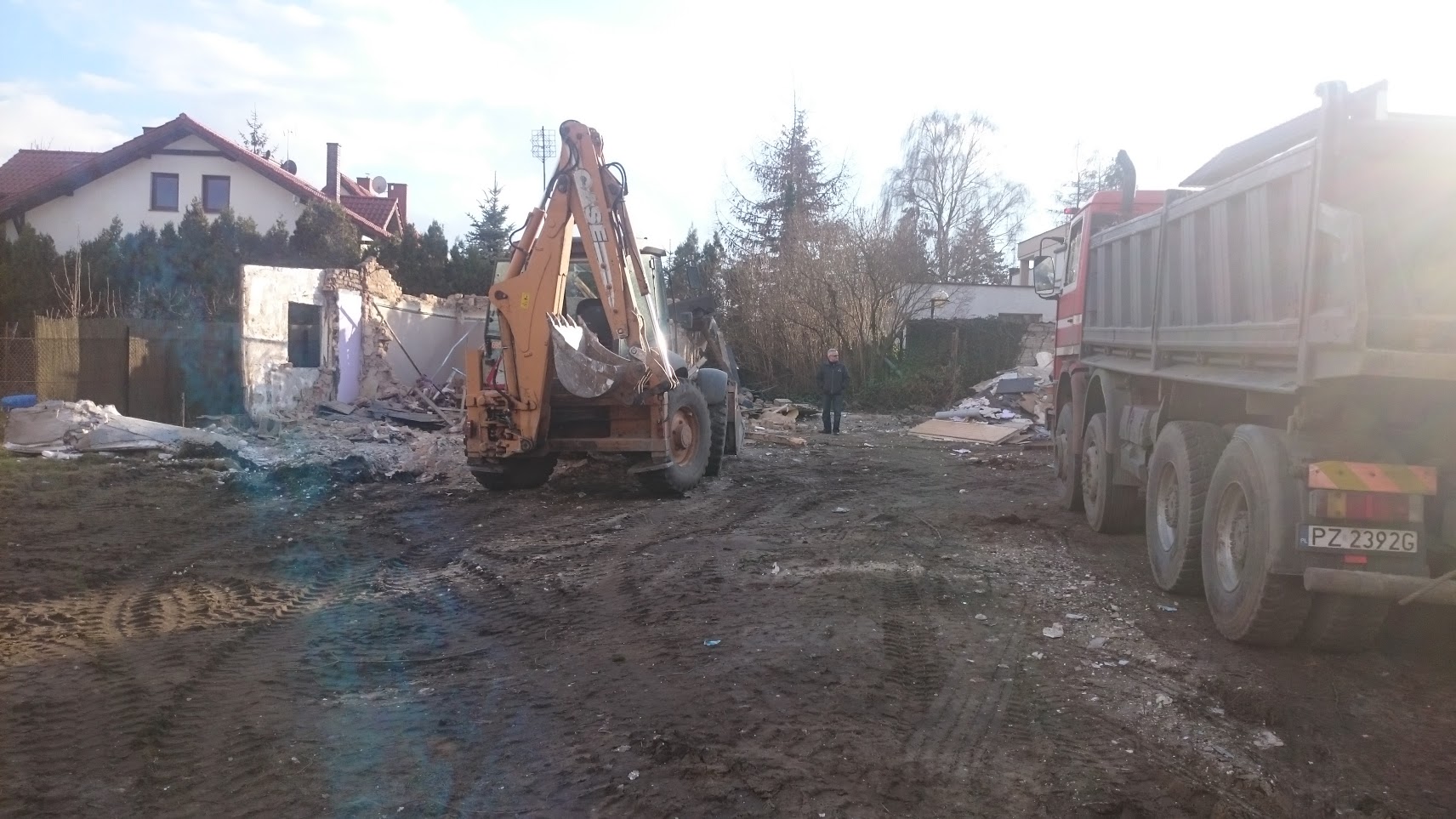 Pomarańczowa koparko-ładowarka Case na placu budowy podczas wyburzania ściany budynku, z gruzem, stojącym mężczyzną i ciężarówką w tle.