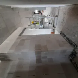 Wnętrze budynku w trakcie budowy, widoczne ściany z białych bloczków oraz rusztowanie z pracownikiem w żółtym kasku w tle.