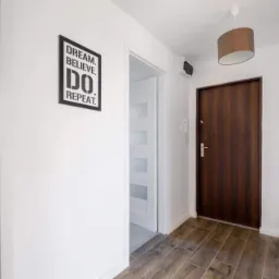 Jasne wnętrze przedpokoju z drewnopodobną podłogą, białymi ścianami, ciemnymi drzwiami wejściowymi i plakatem motywacyjnym 'Dream. Believe. Do. Repeat.'