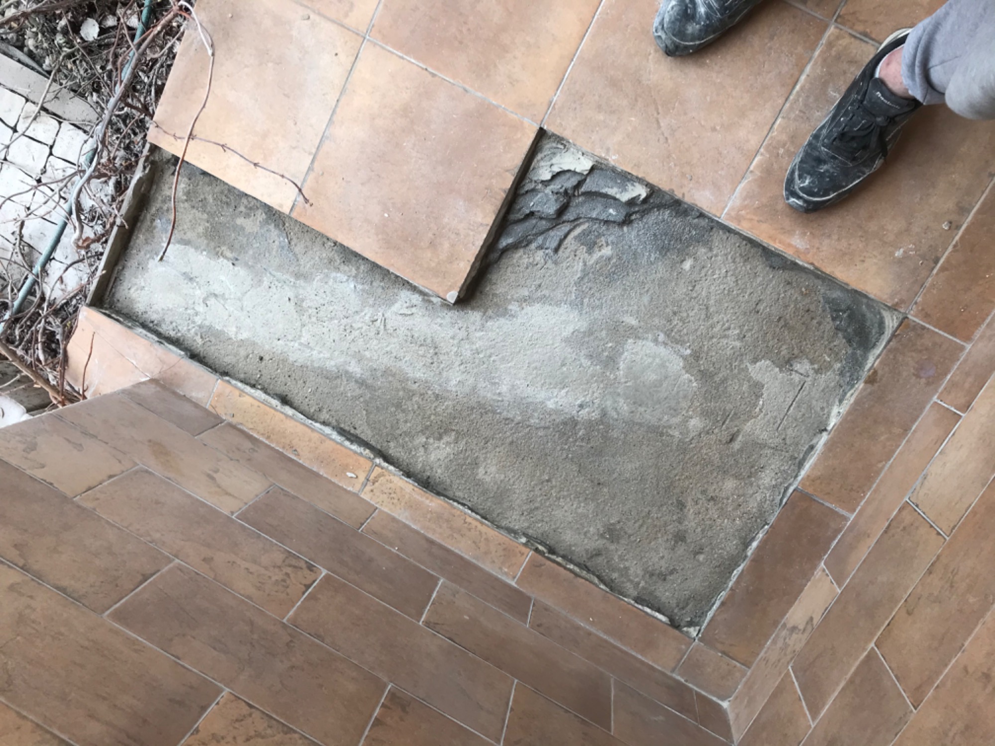 Fragment podłogi z brązowych płytek w trakcie naprawy w Warszawie, widoczny ubytek i przygotowane podłoże pod nową płytkę, buty robocze w tle.