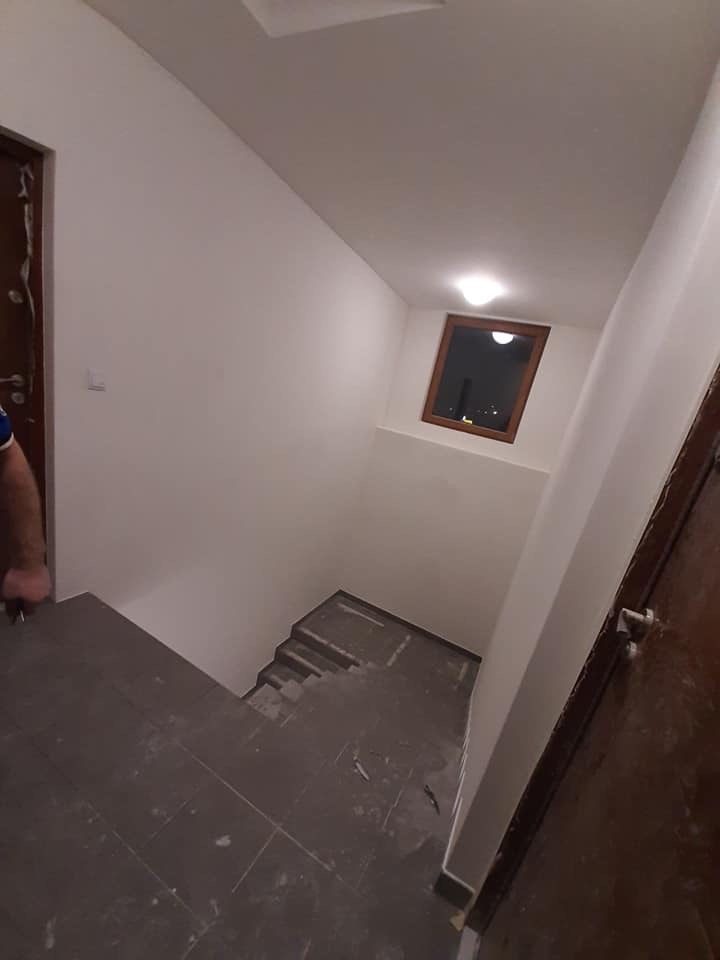 Klatka schodowa w trakcie remontu: świeżo pomalowane ściany na biało, szare płytki na schodach, drewniane okno z widokiem na nocne miasto.