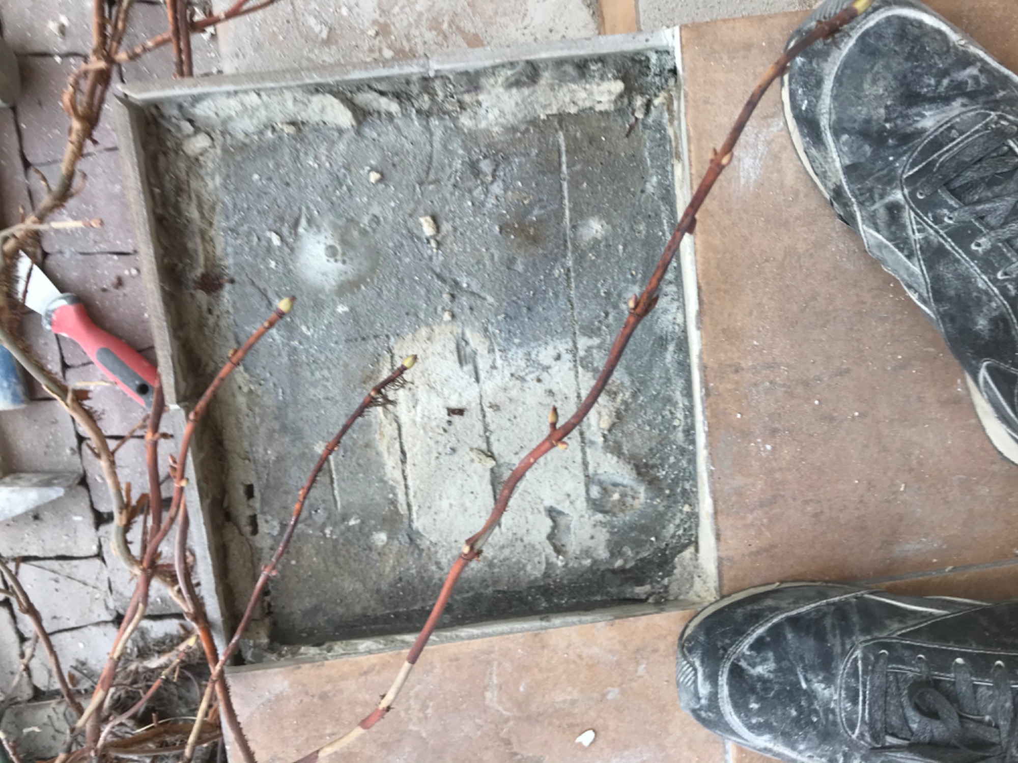 Odkryta, kwadratowa przestrzeń w podłodze z resztkami betonu i widocznymi warstwami podłoża, obok leżące gałązki i but roboczy.