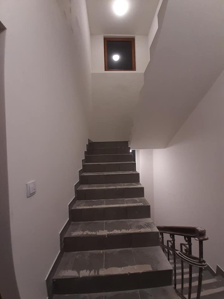 Klatka schodowa w jasnych barwach z ciemnymi, wykafelkowanymi schodami i drewnianą balustradą, oświetlona górnym światłem i małym okienkiem.