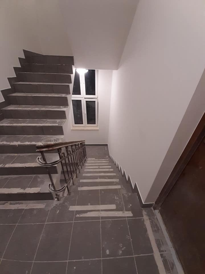 Klatka schodowa w trakcie odświeżania, widoczne zabezpieczenia na schodach i świeżo pomalowane ściany, ciemne płytki podłogowe i metalowa balustrada.