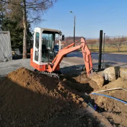 Mini koparka w trakcie wykopu ziemi, widoczne niebieskie rury instalacyjne w wykopie, obok sterta ziemi i kontener biurowy w tle.