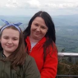 Dwie uśmiechnięte kobiety na tle górskiego krajobrazu, widoczne z platformy widokowej. Jedna w czerwonej bluzie, druga w zielonej kurtce z niebieską opaską.